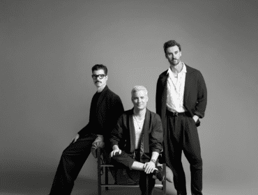 Three men in stylish black suits posing against a grey background.